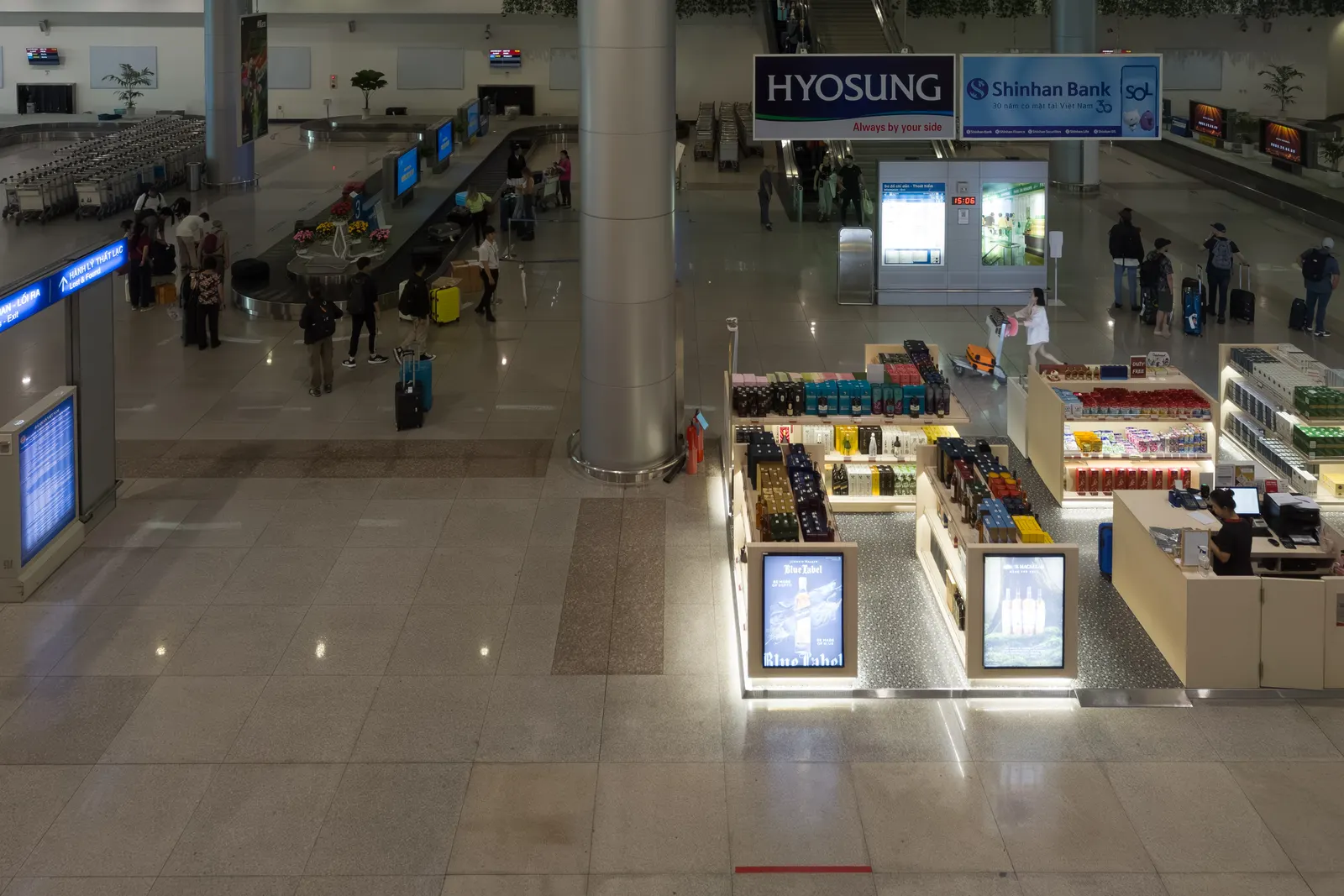 Overhead view of an airport arrivals hall with a duty-free kiosk, advertising signs and travellers with luggage.
