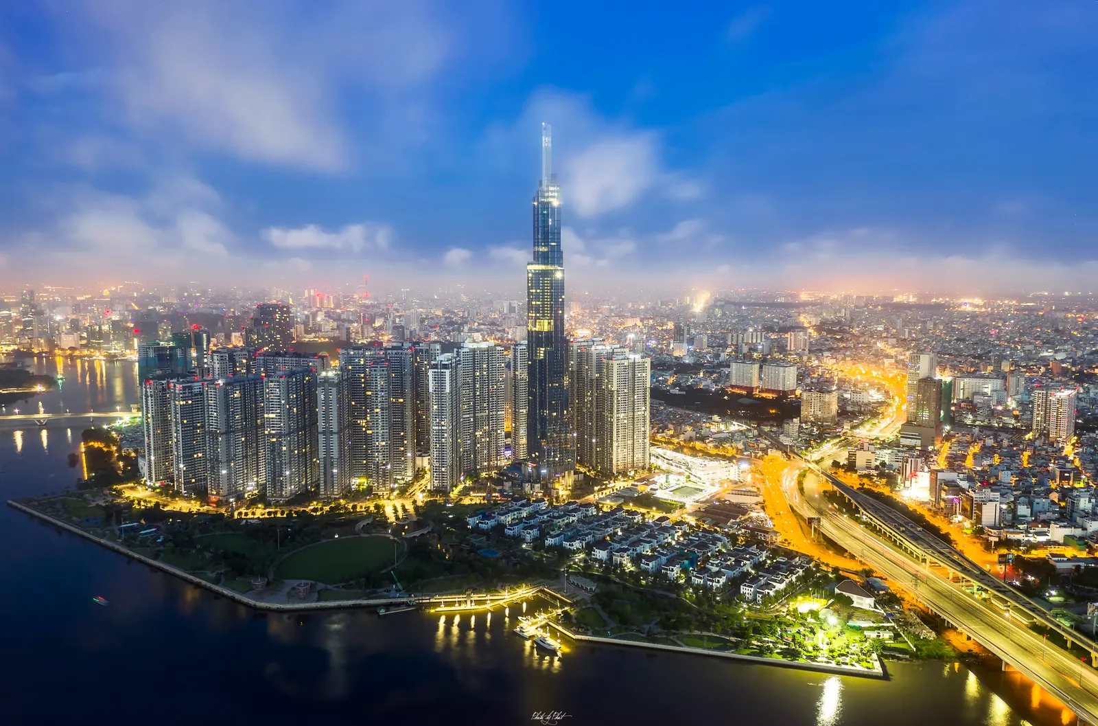 Night view of a very tall tower above a lit riverside park and a curving expressway.