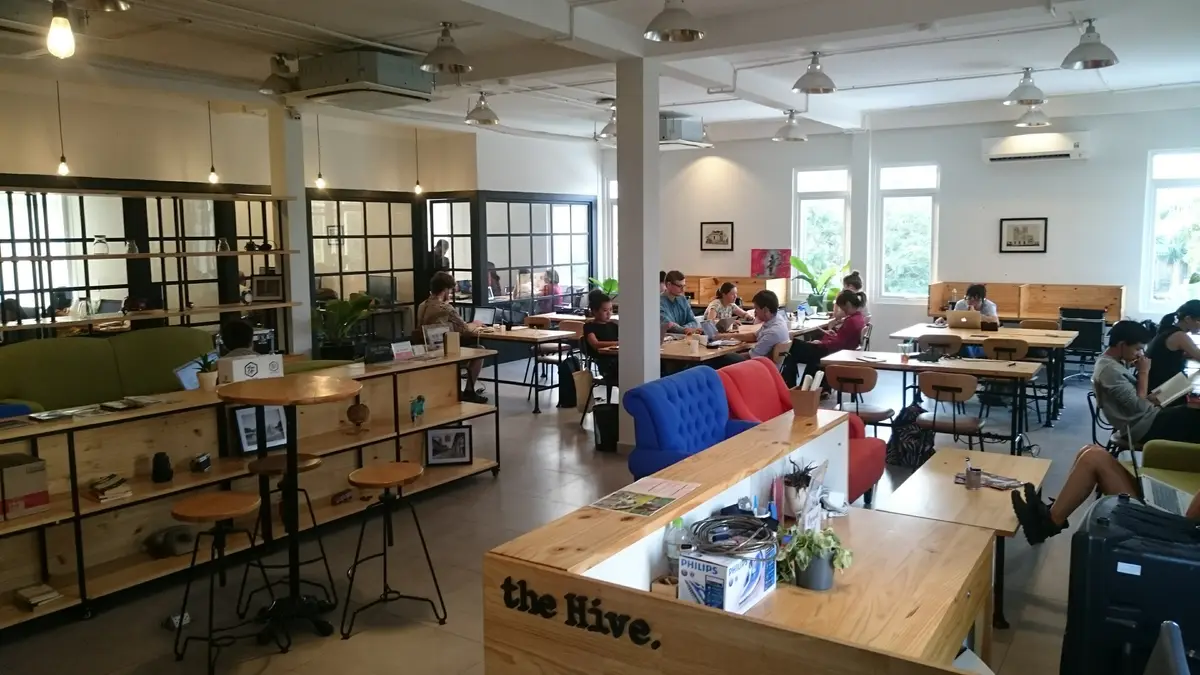 Open-plan coworking room with wooden desks, stools and people working under ceiling fans