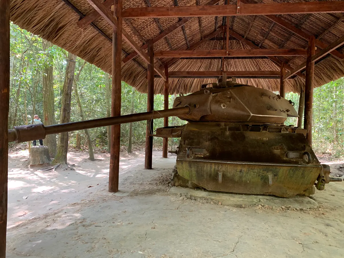 Rusted tank with its gun barrel under a thatched shelter in a forest