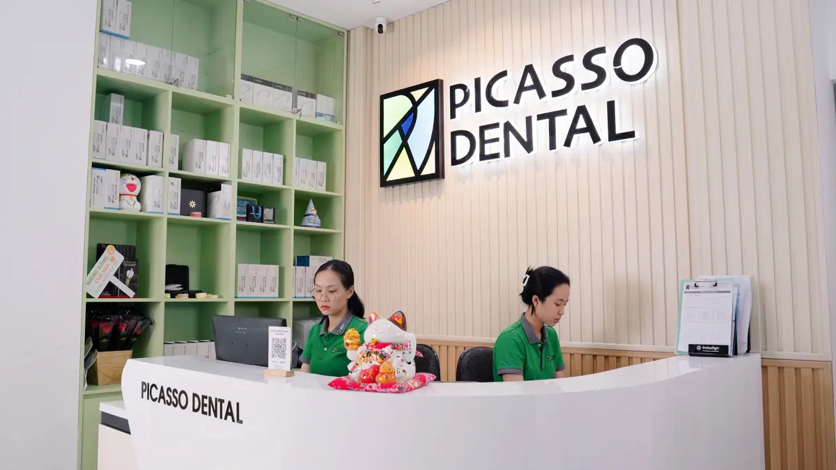 Reception desk with the Picasso Dental logo on a wooden wall and shelves of dental products behind two staff.