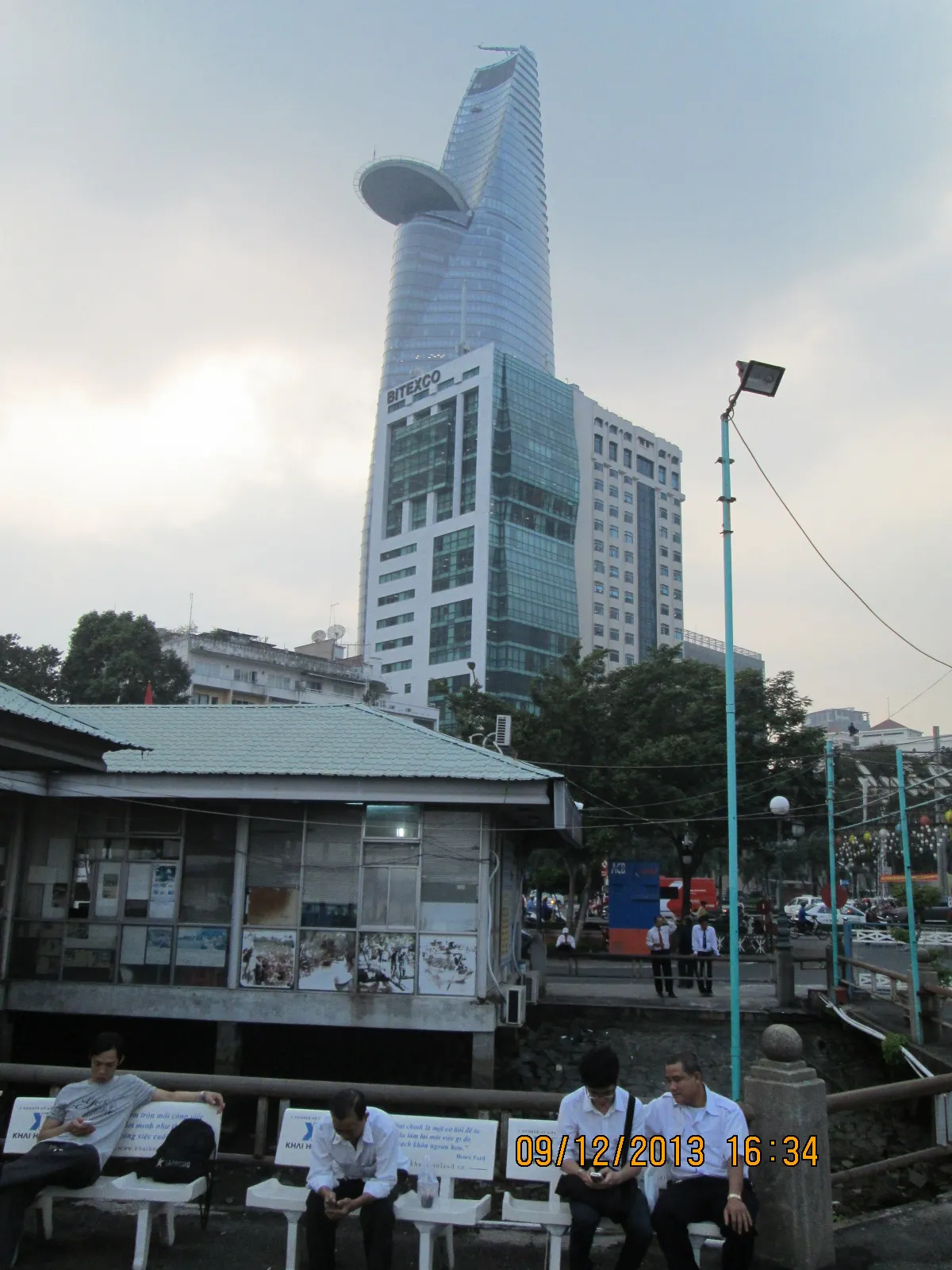 Glass office tower rising behind trees and a small kiosk on a riverside street