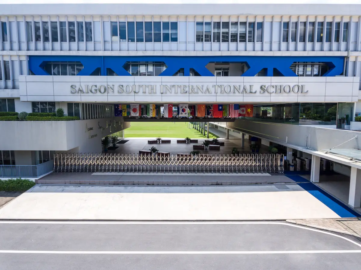 Glass-fronted school building with a blue band and the school name above a paved forecourt