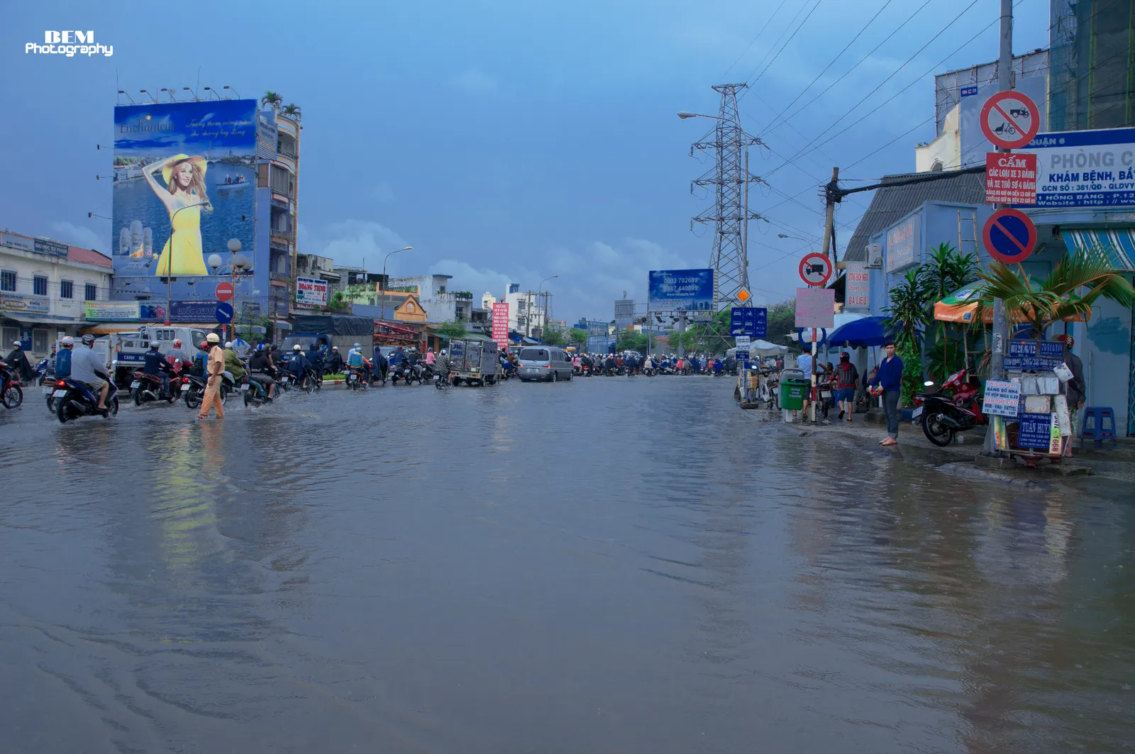 Wide street under a blue evening sky with water covering the road and motorbikes and pedestrians wading in the distance