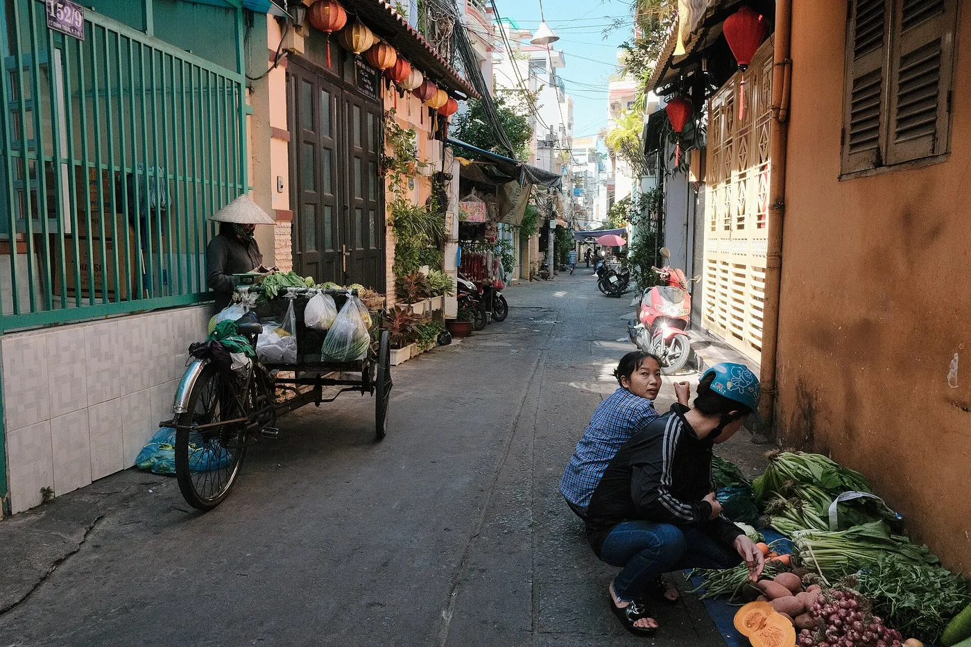 Narrow paved alley between houses in Ho Chi Minh City with a vendor tricycle and a woman sorting vegetables