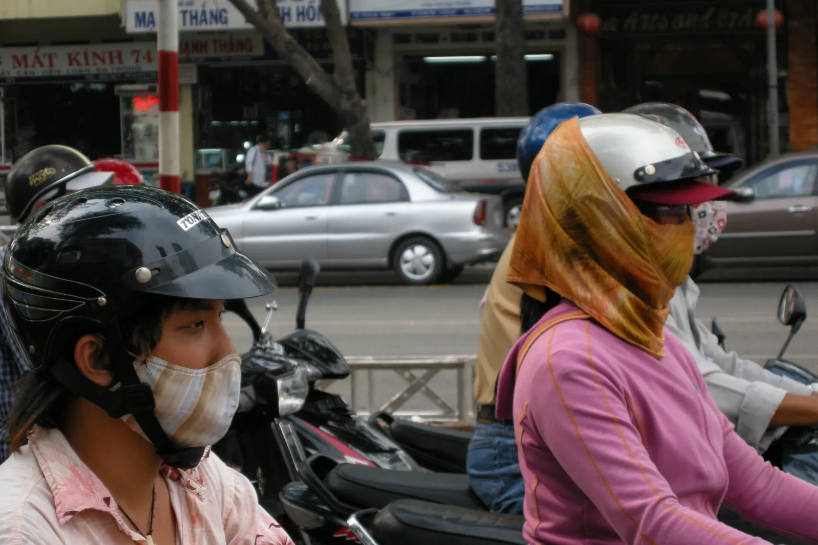 Motorbike riders in helmets and face masks at a street junction with a car behind