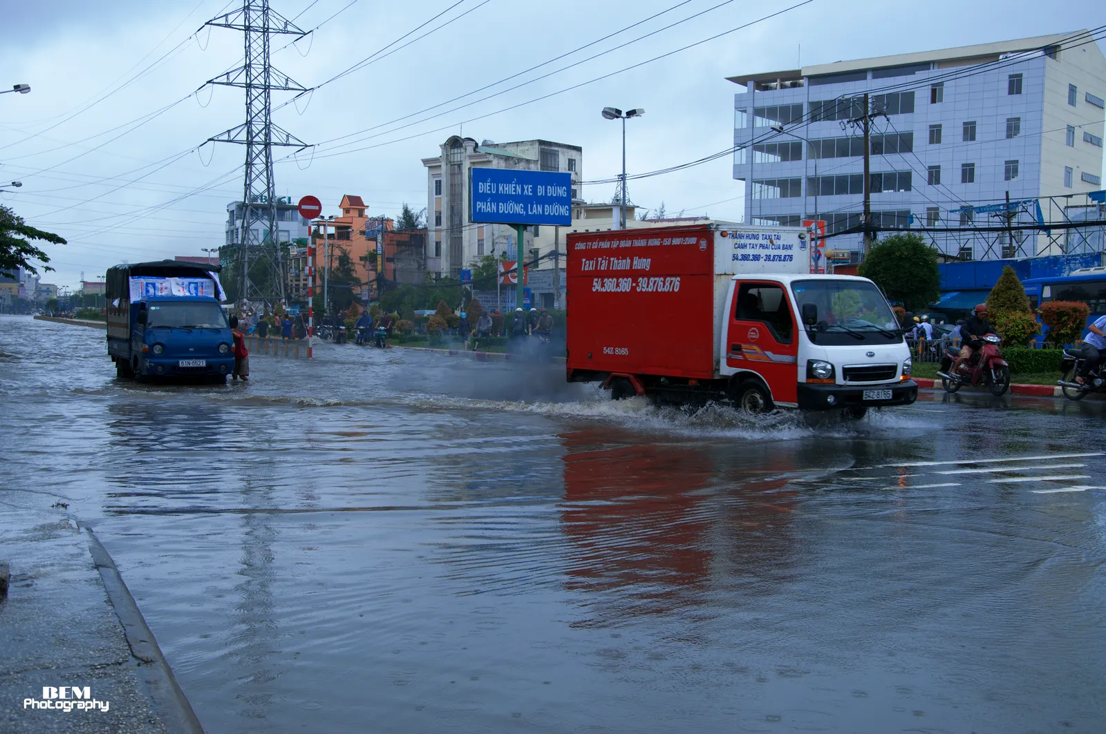 Delivery trucks driving through shallow water on a wide road with pylons in the distance