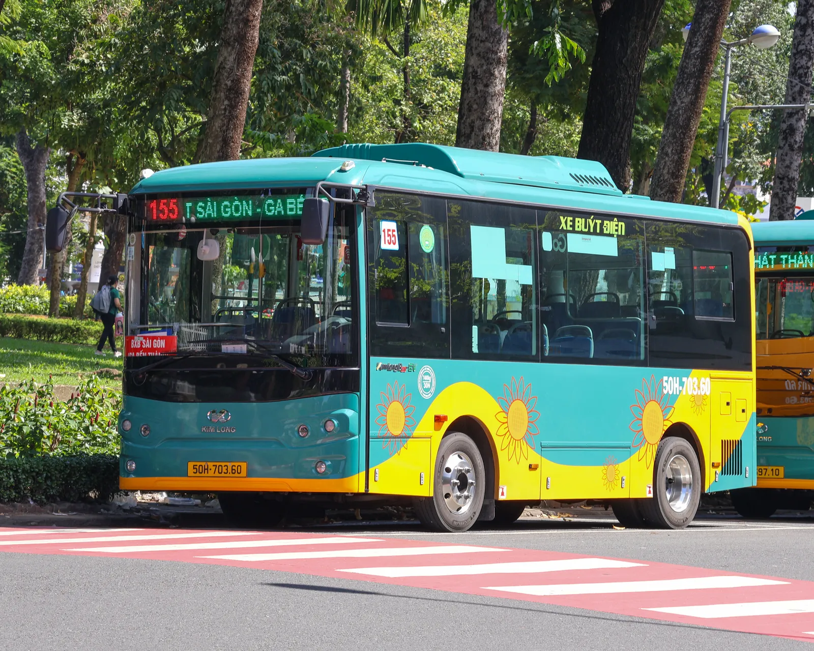 Teal and yellow city bus with a lit destination sign beside a tree-lined park