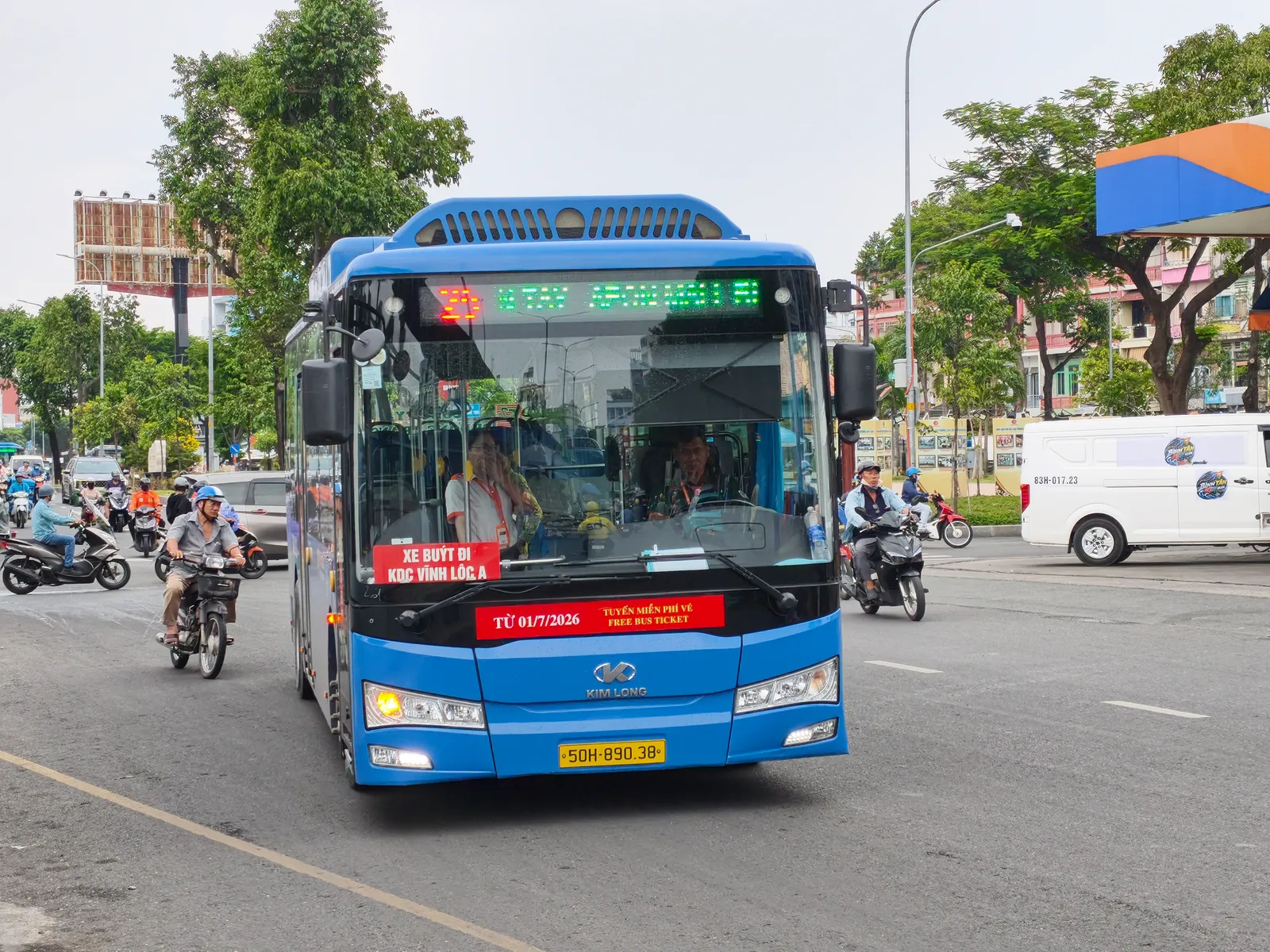 Blue city bus with a red sign in the windscreen on a road with motorbikes and a white van