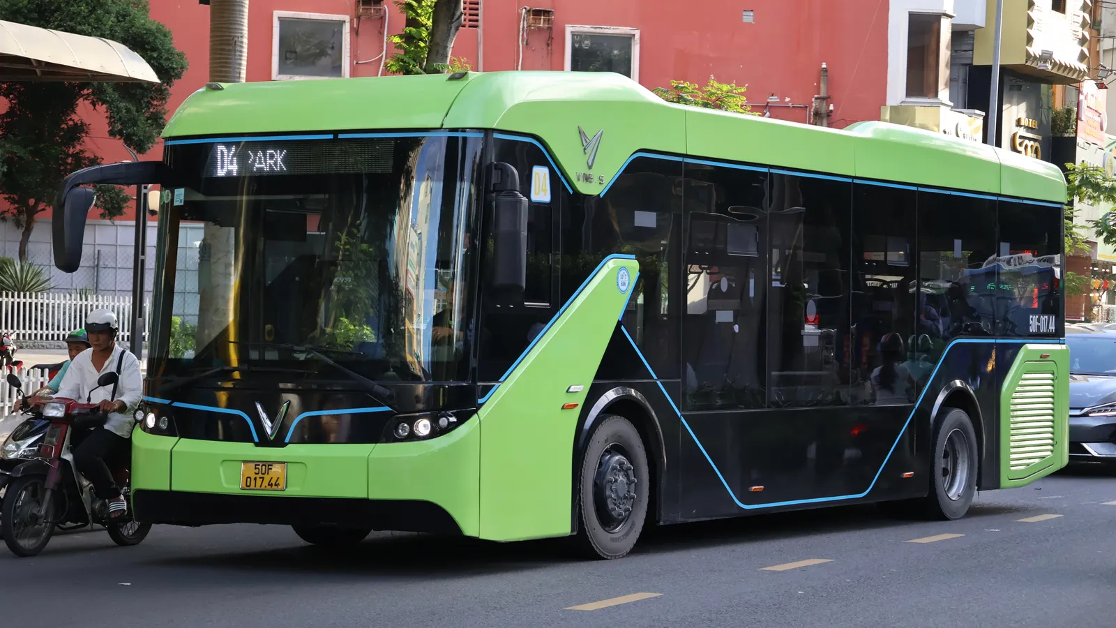 Green and black electric bus with a lit route sign stopped on a street beside a person on a motorbike