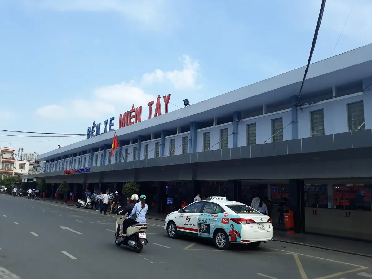 Long low station building with the Ben Xe Mien Tay sign above a street where a white taxi passes
