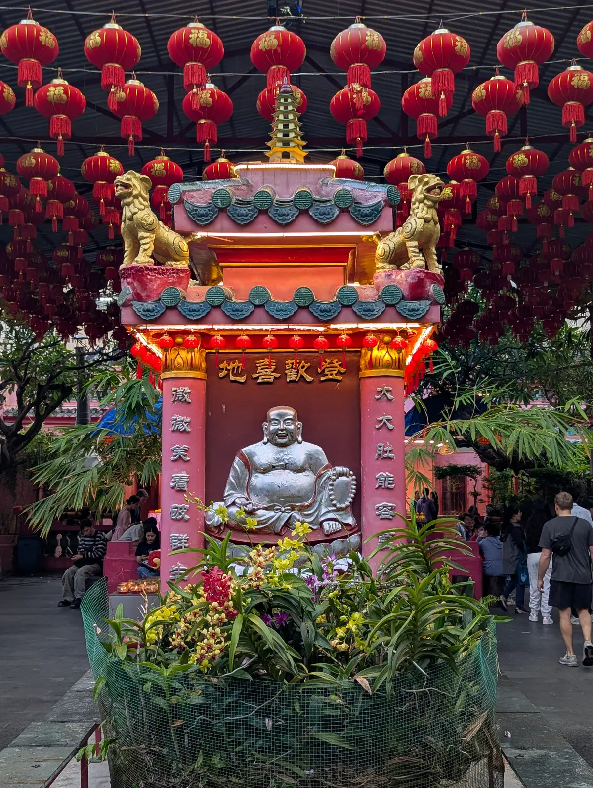White laughing Buddha statue in a pink and red shrine topped by two golden lions, with plants around it