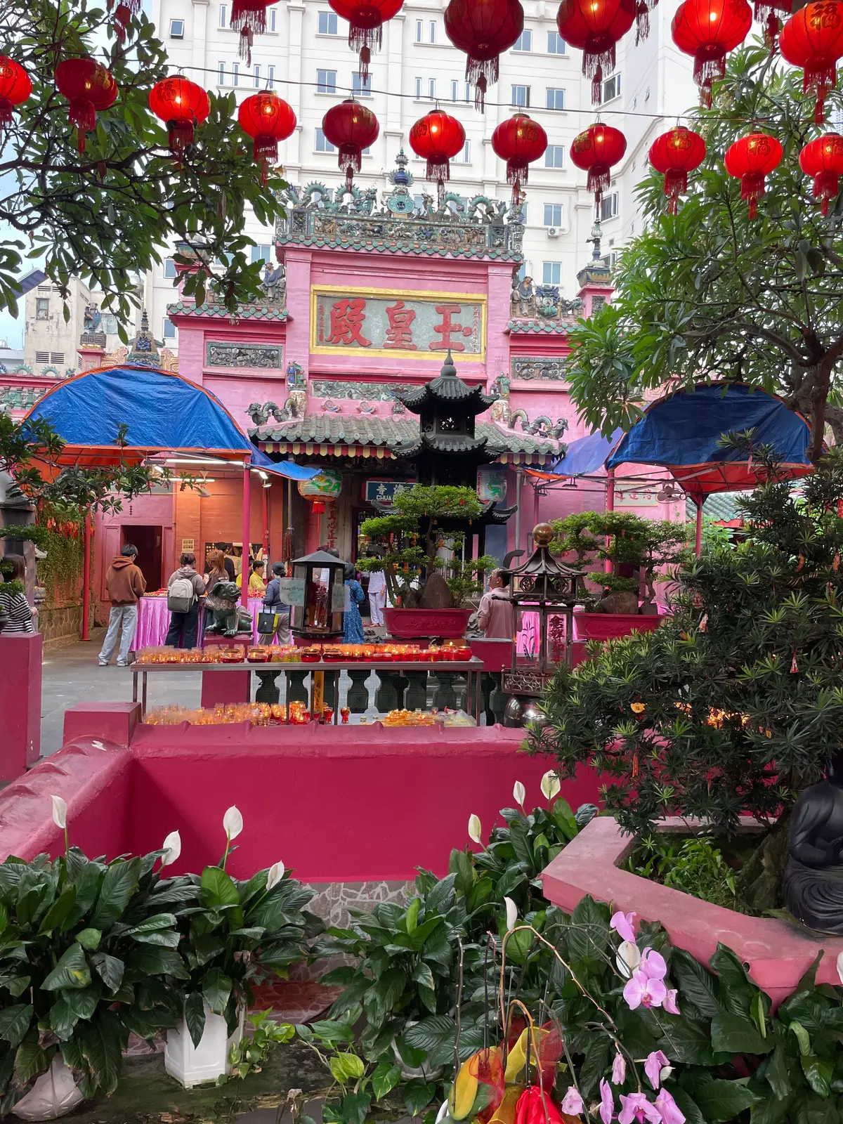 Courtyard with pink walls, a red offering table, a tiered incense burner and visitors under blue tarpaulins