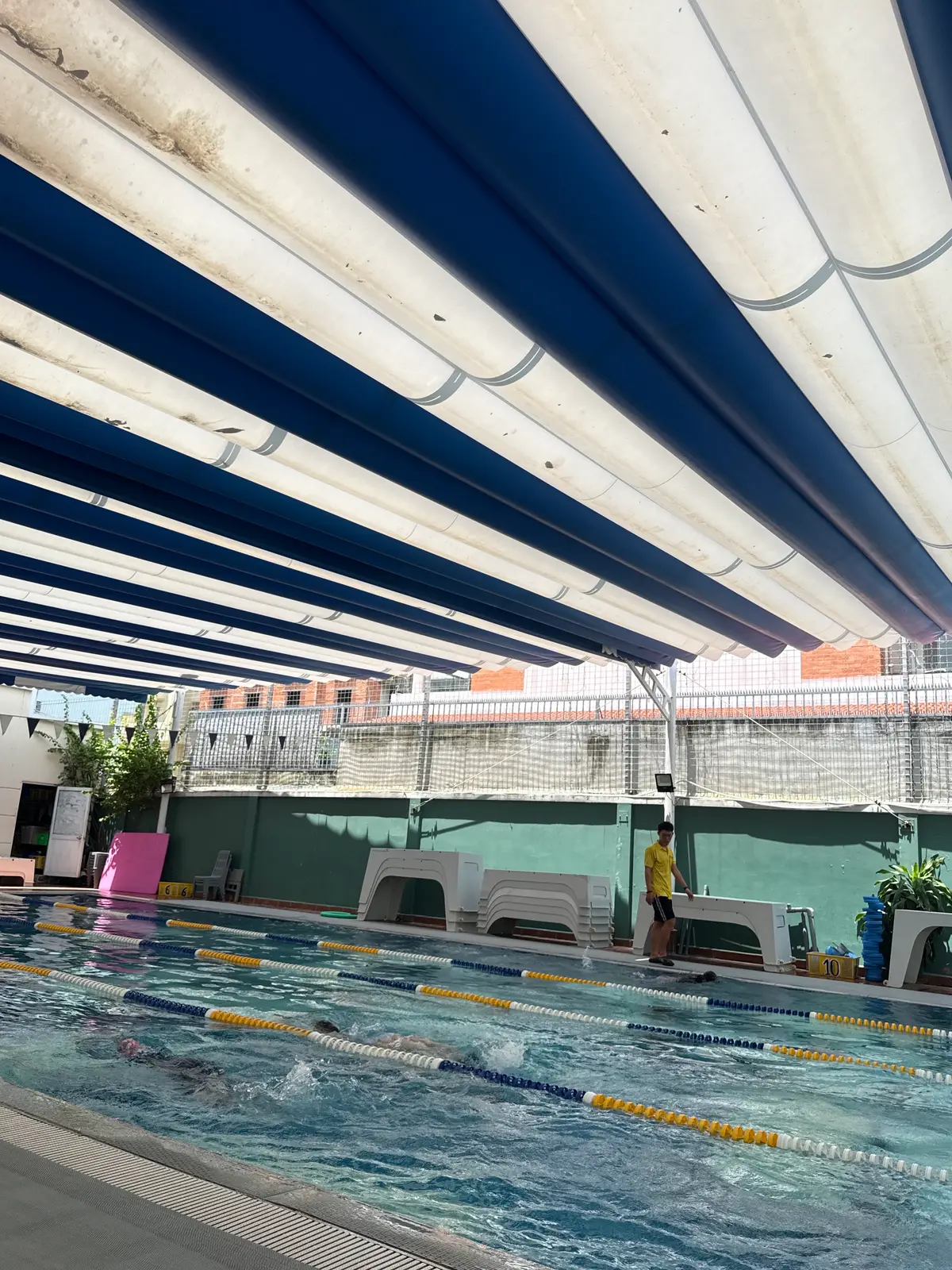 Pool deck with blue shade sails over white loungers beside a green wall