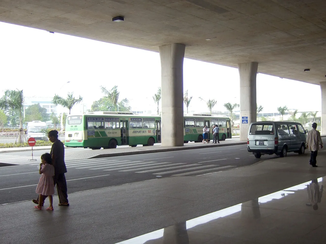 Green city buses parked under a covered airport forecourt with a minivan and a few people walking