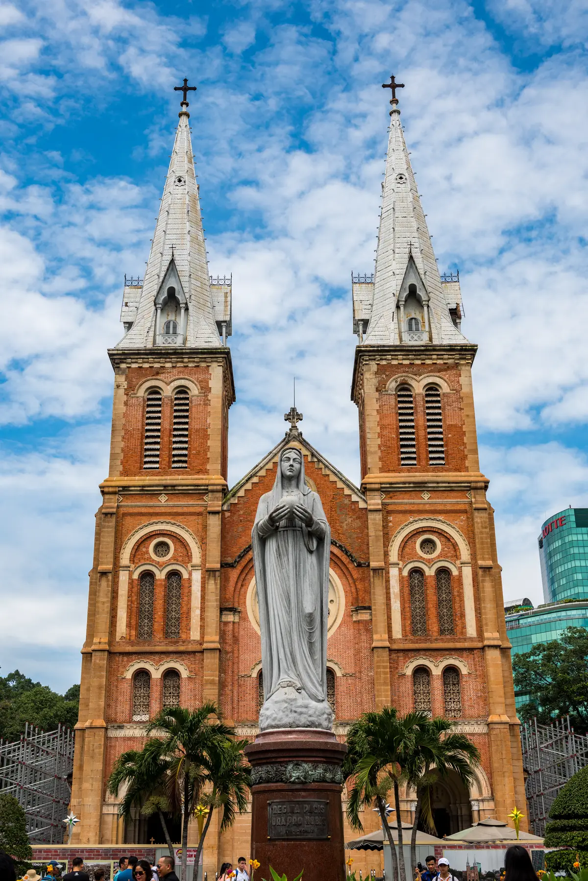 Two red-brick bell towers with arched windows behind a white statue of the Virgin Mary under a blue sky