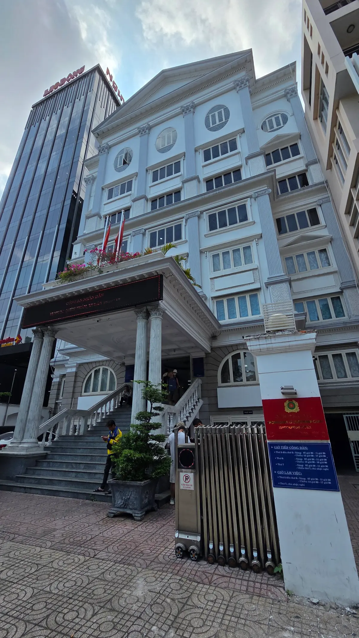 Front of a white multi-storey office building with columns, a red police crest and flags on the facade, seen from the pavement.