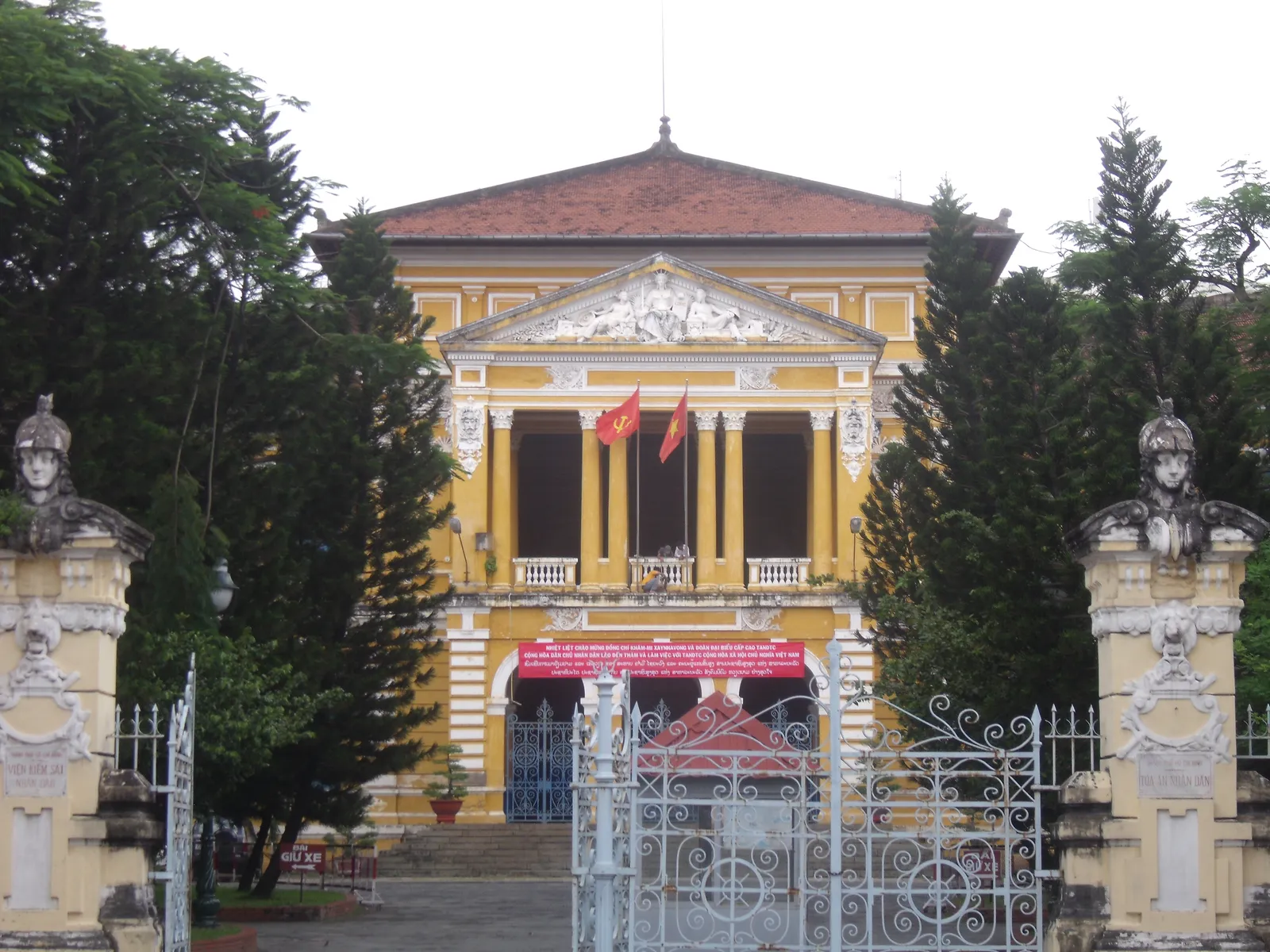 Yellow colonial-style building with a columned porch and red roof behind an iron gate and trees