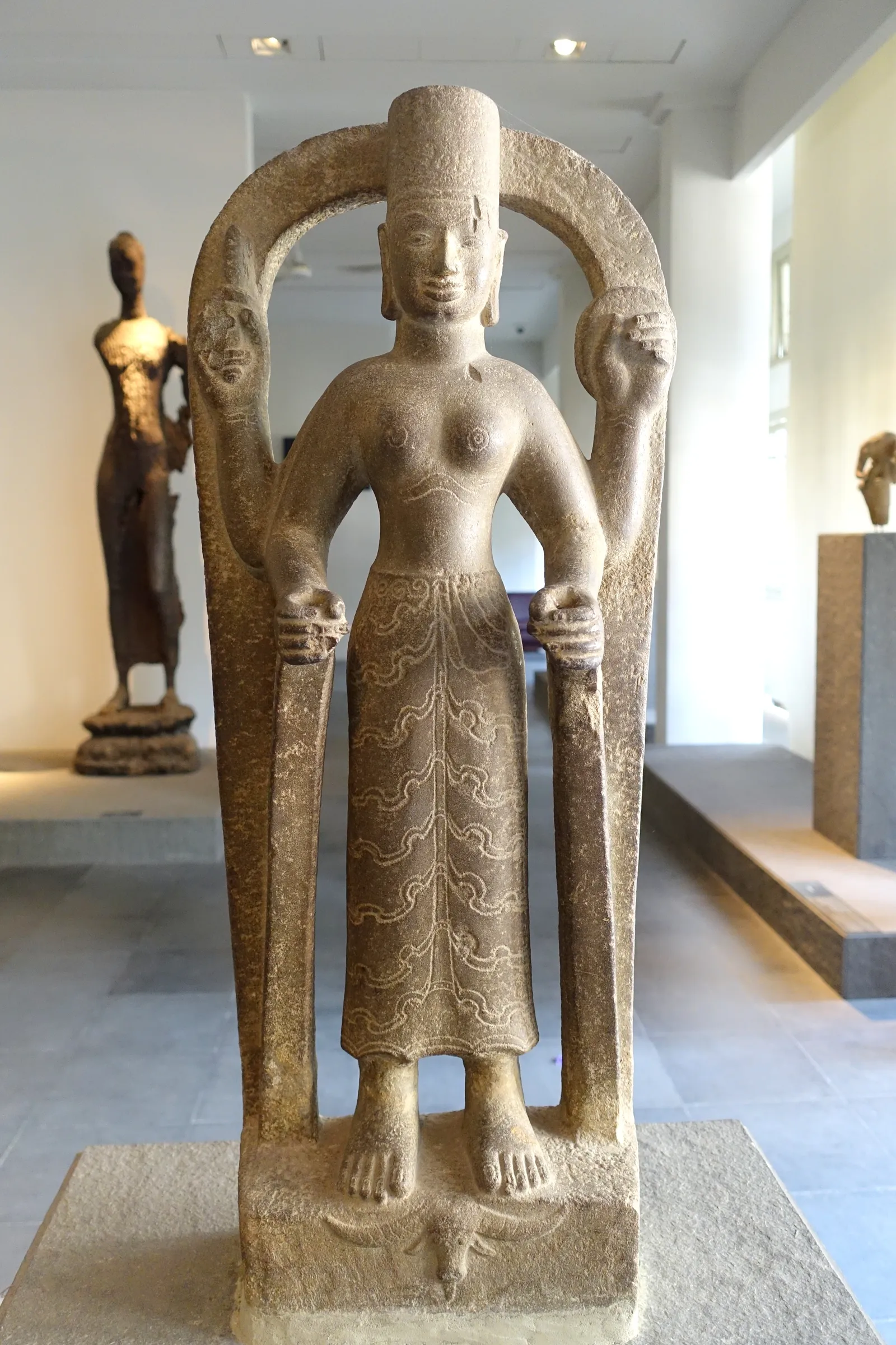 Tall carved stone goddess statue standing inside a pale arched frame in a gallery
