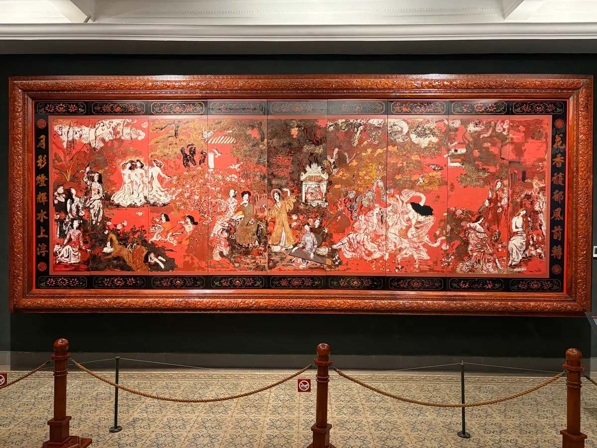 Large framed lacquer painting with red and gold figures behind a rope barrier in a gallery