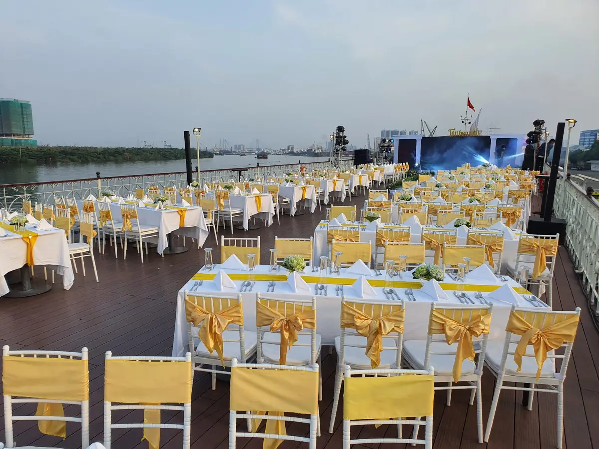 Open upper deck set with long tables, white chairs and yellow sashes beside the river at dusk