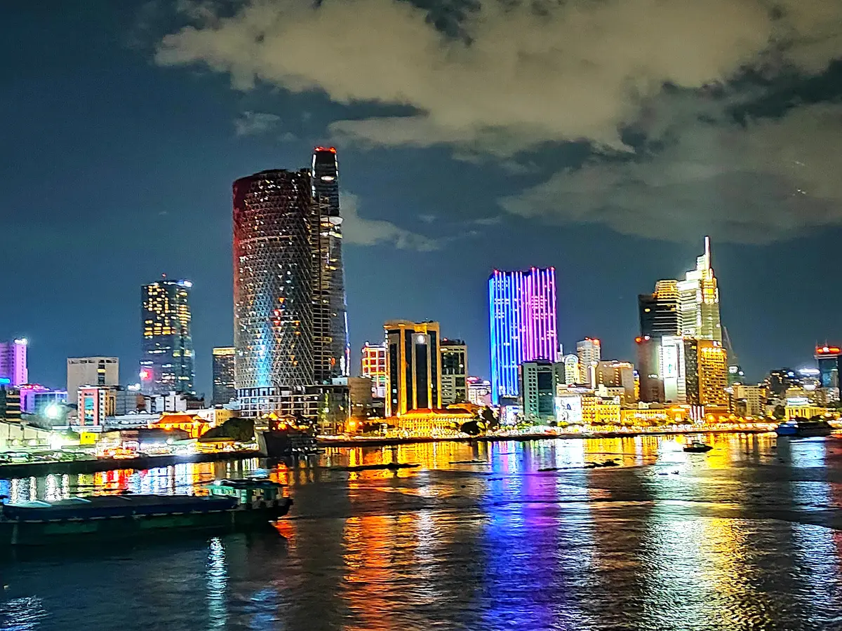City skyline across the river at night with boats moored in the foreground and colourful lights on the water