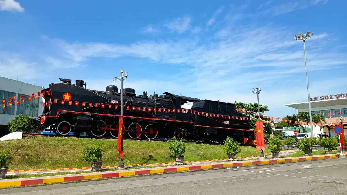 Black locomotive on display on a grass verge beside a road under a blue sky