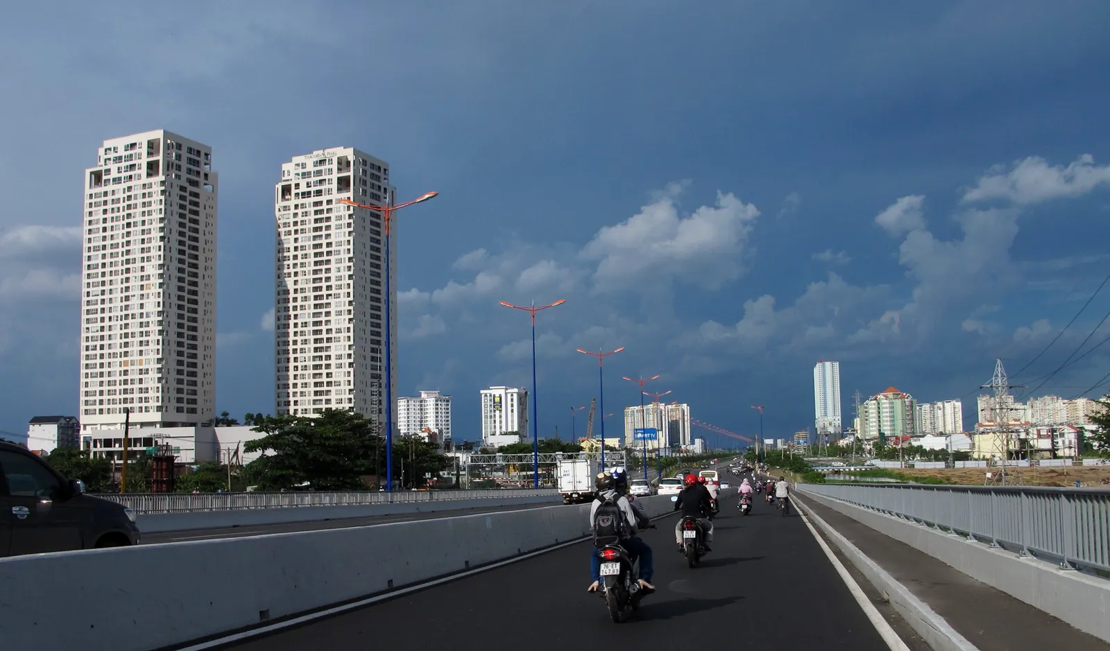 Two tall apartment towers beside a busy road with motorbikes under a dark blue sky