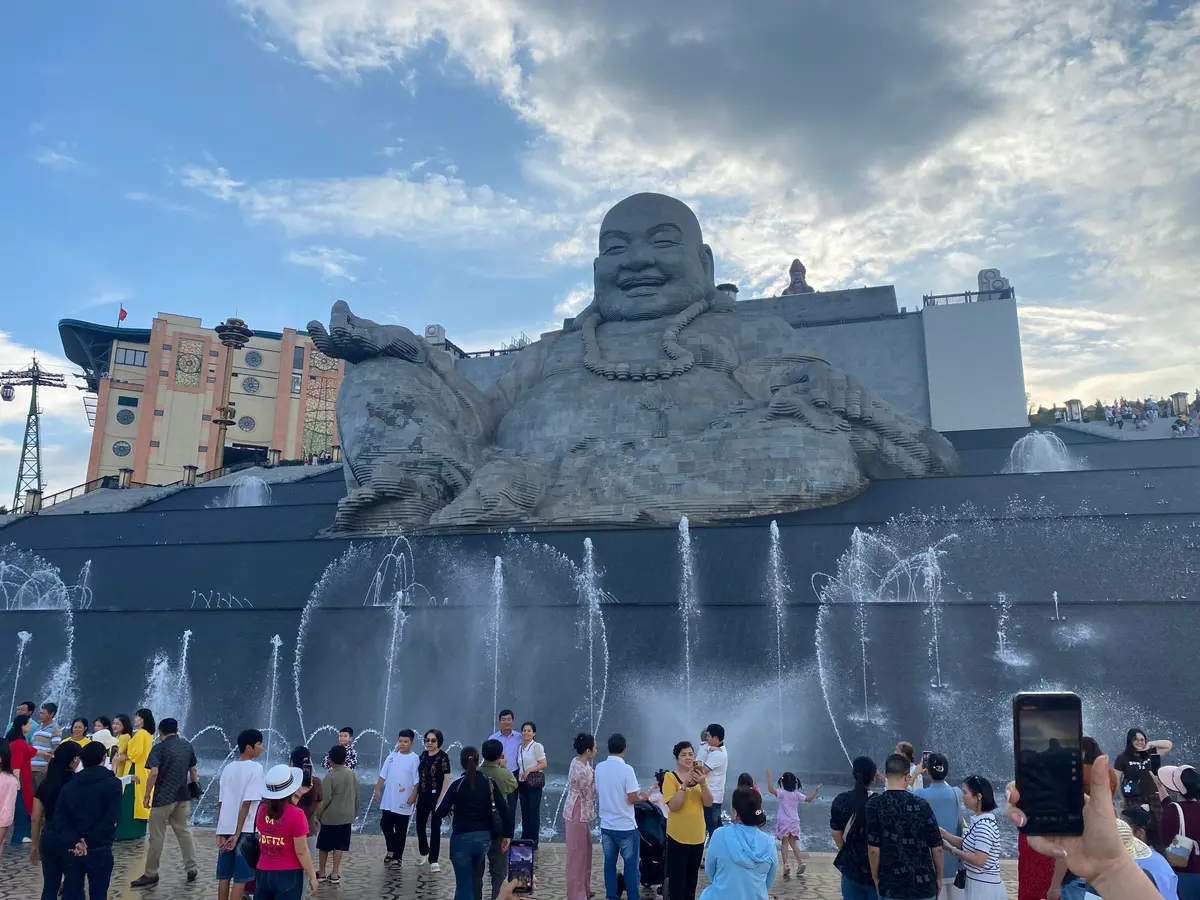 Large stone laughing Buddha statue behind a fountain with a crowd of visitors in front