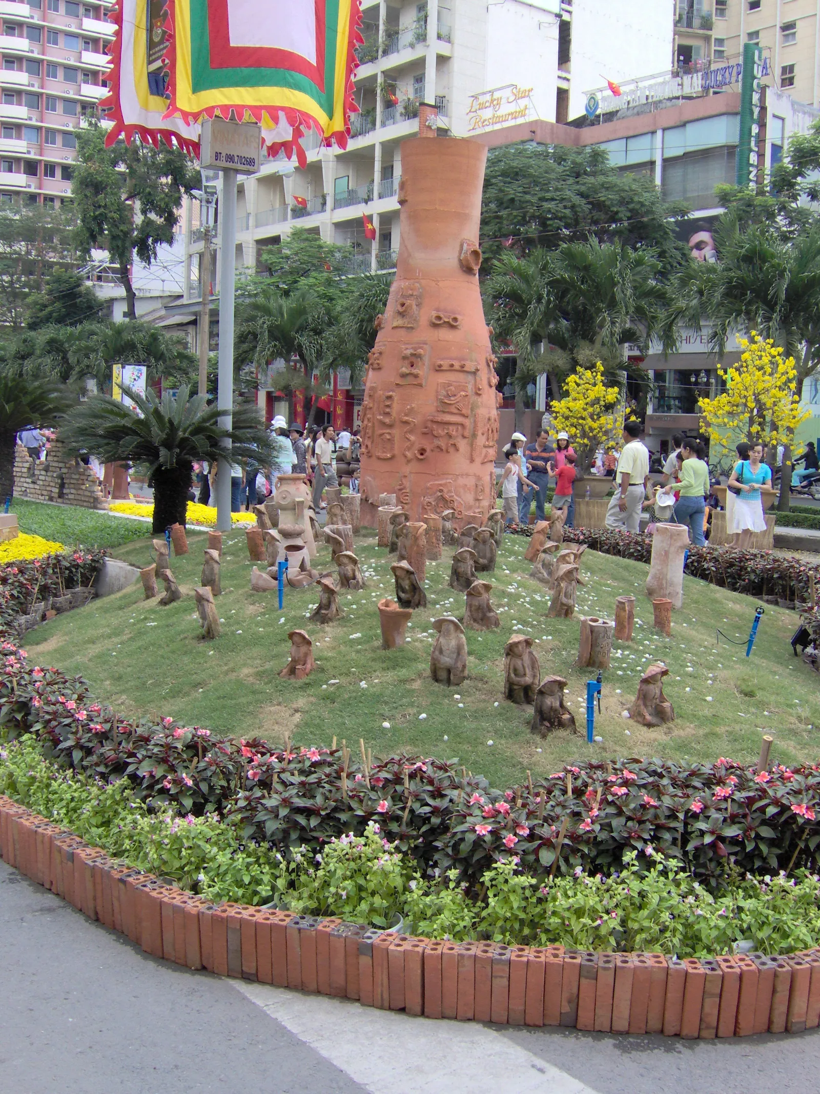 Round flower bed with small clay figures and a tall pottery vase on a city corner
