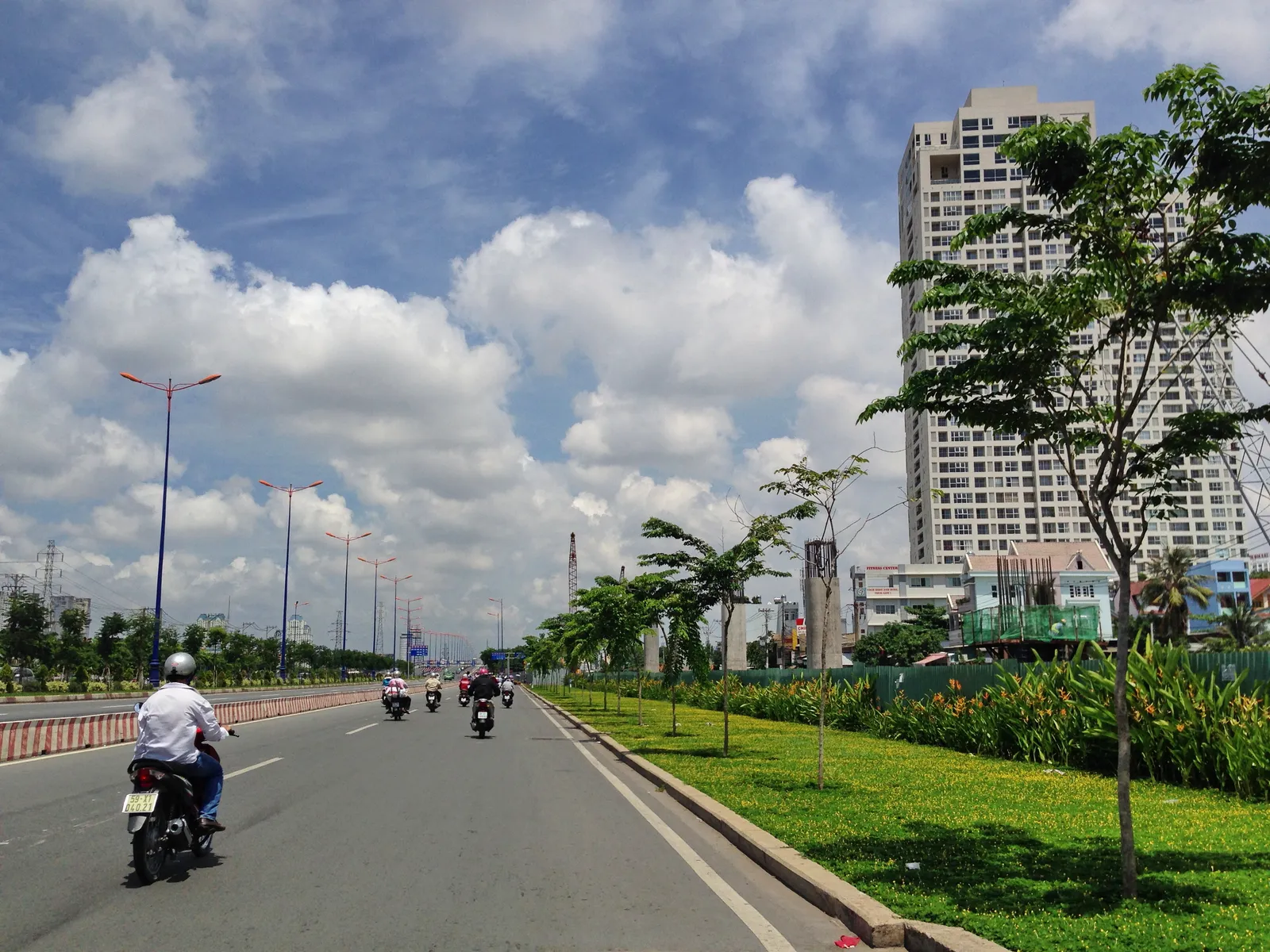 Wide road with motorbikes beside a green median and tall apartment towers in the distance