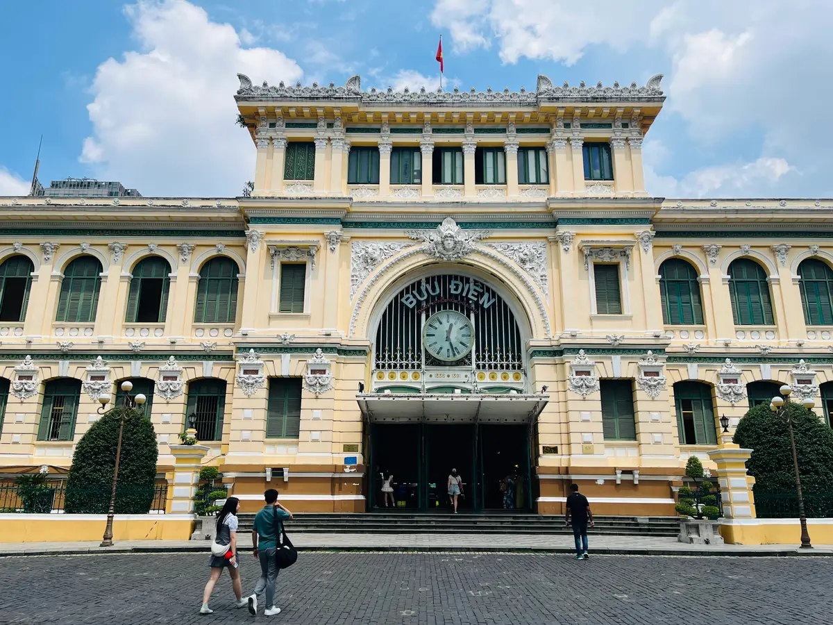 Yellow colonial building with a large clock above the arched entrance and visitors crossing the square