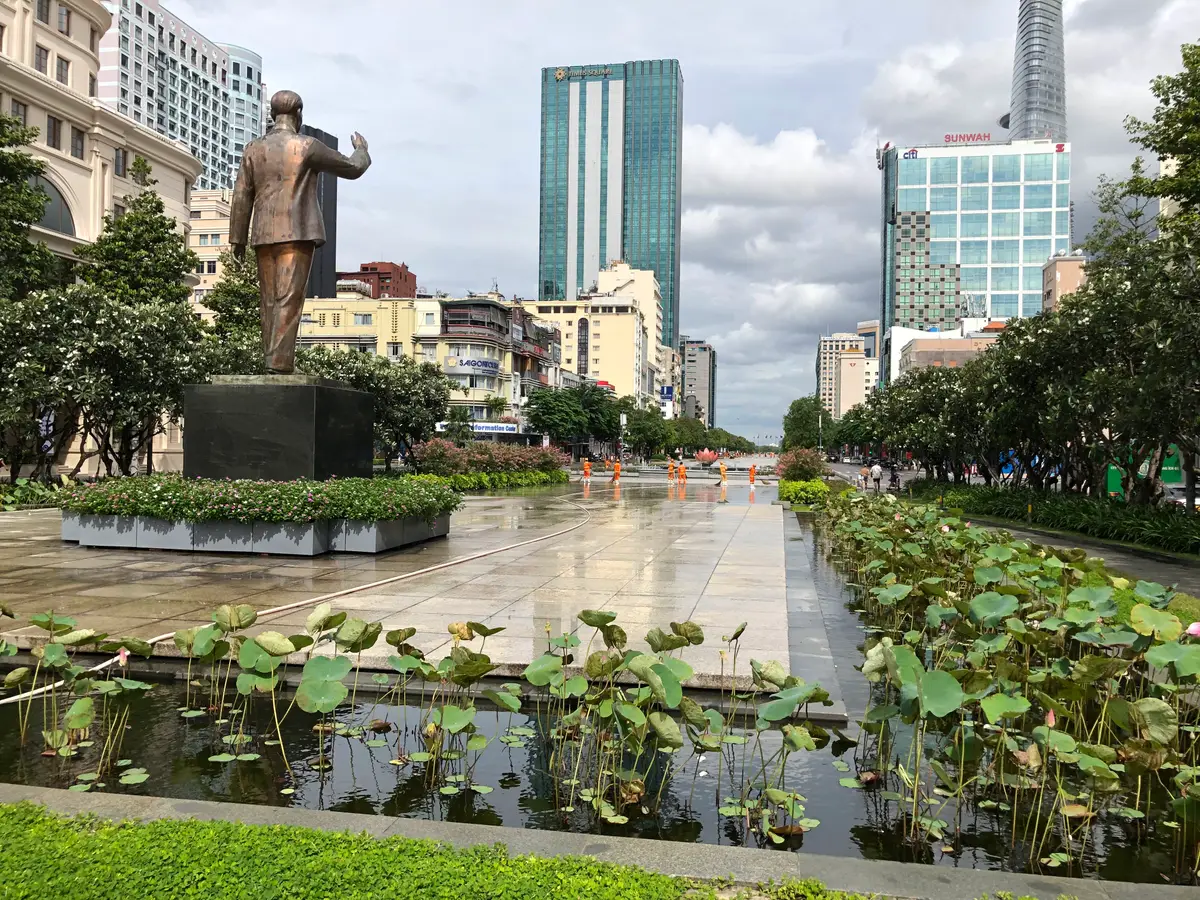 Wide wet pavement with a lotus pond on the right, a bronze statue on the left and office towers behind