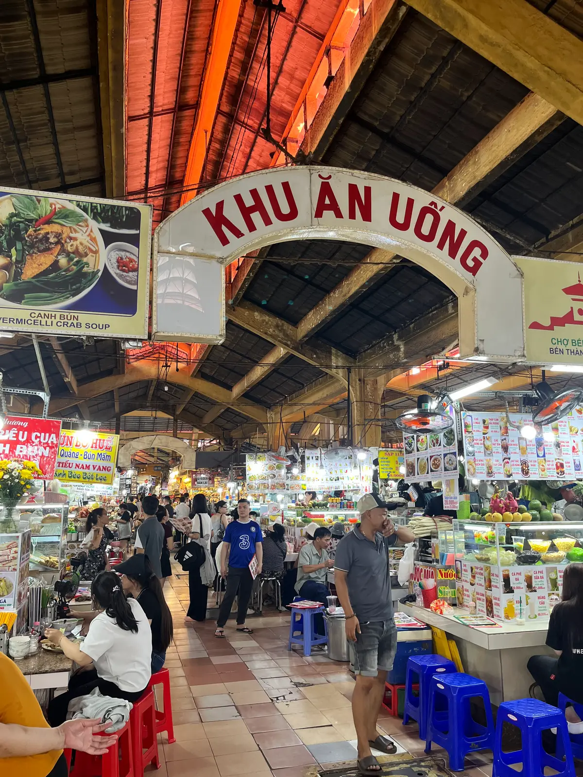 Crowded food court under a metal roof with an overhead sign and rows of stalls