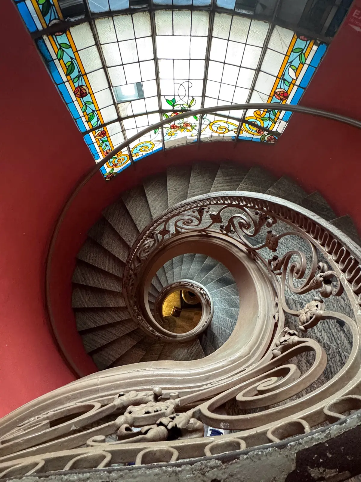 Spiral staircase with an ornate iron railing seen from above beneath a stained glass skylight