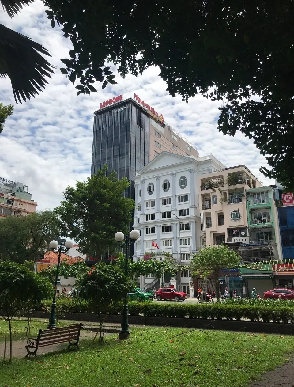 Park bench and trees in the foreground with a white multi-storey building and a tall glass tower behind them.