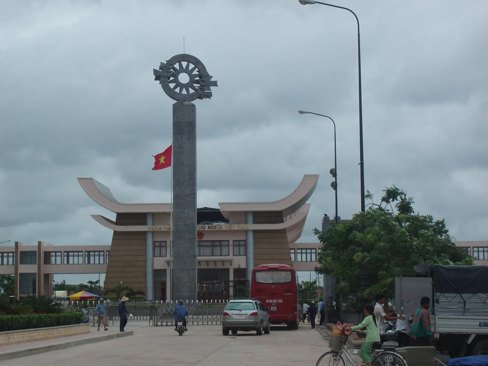 Road at the Moc Bai border gate with a red bus, a tall monument with a red flag and the low border building behind it under a grey sky