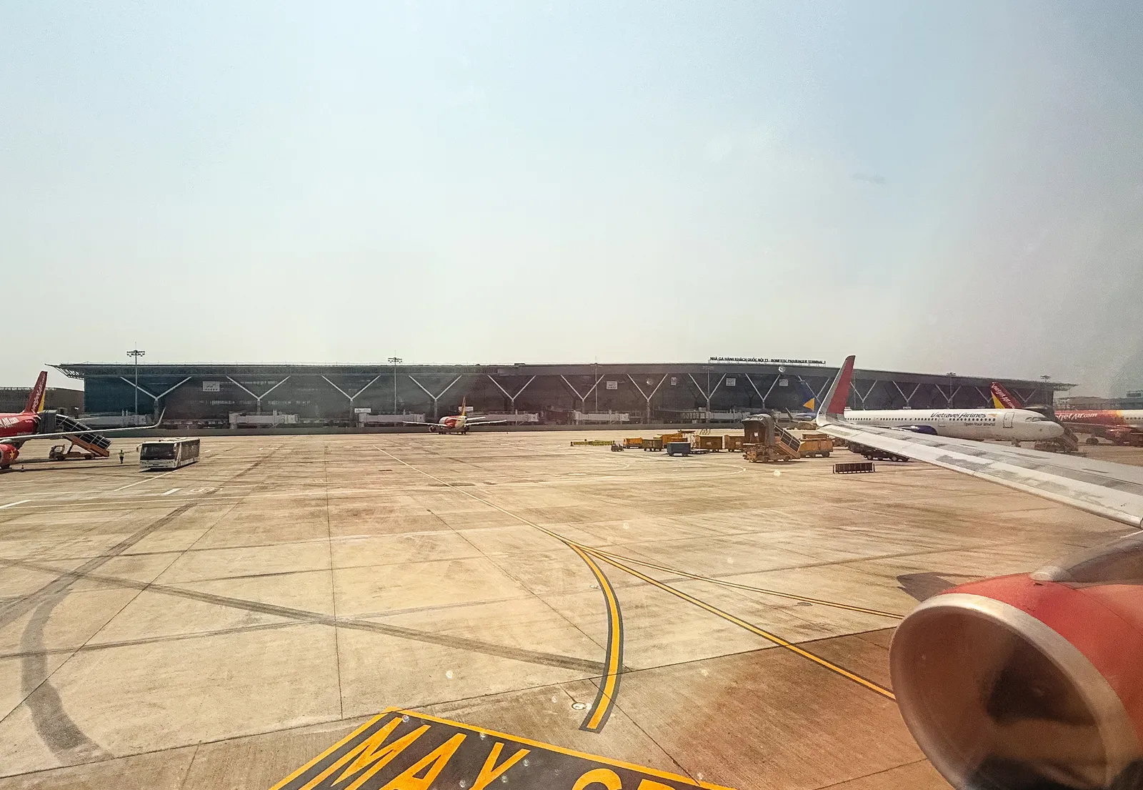 Hazy view across the Tan Son Nhat apron from an aircraft window, with a long terminal building and painted markings on the ground