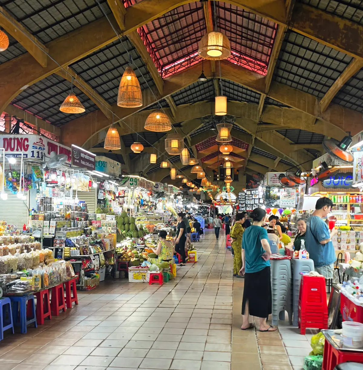 Covered market aisle with fruit stalls under a wooden roof hung with round lamps